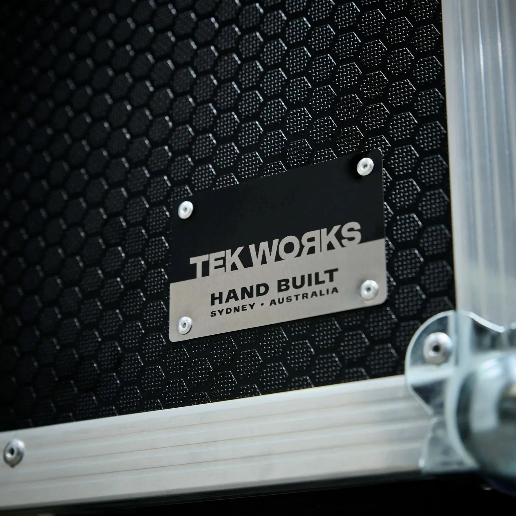 Close-up of a black textured surface with a metallic nameplate reading 'TEK WORKS HAND BUILT SYDNEY AUSTRALIA' and part of a metallic frame.