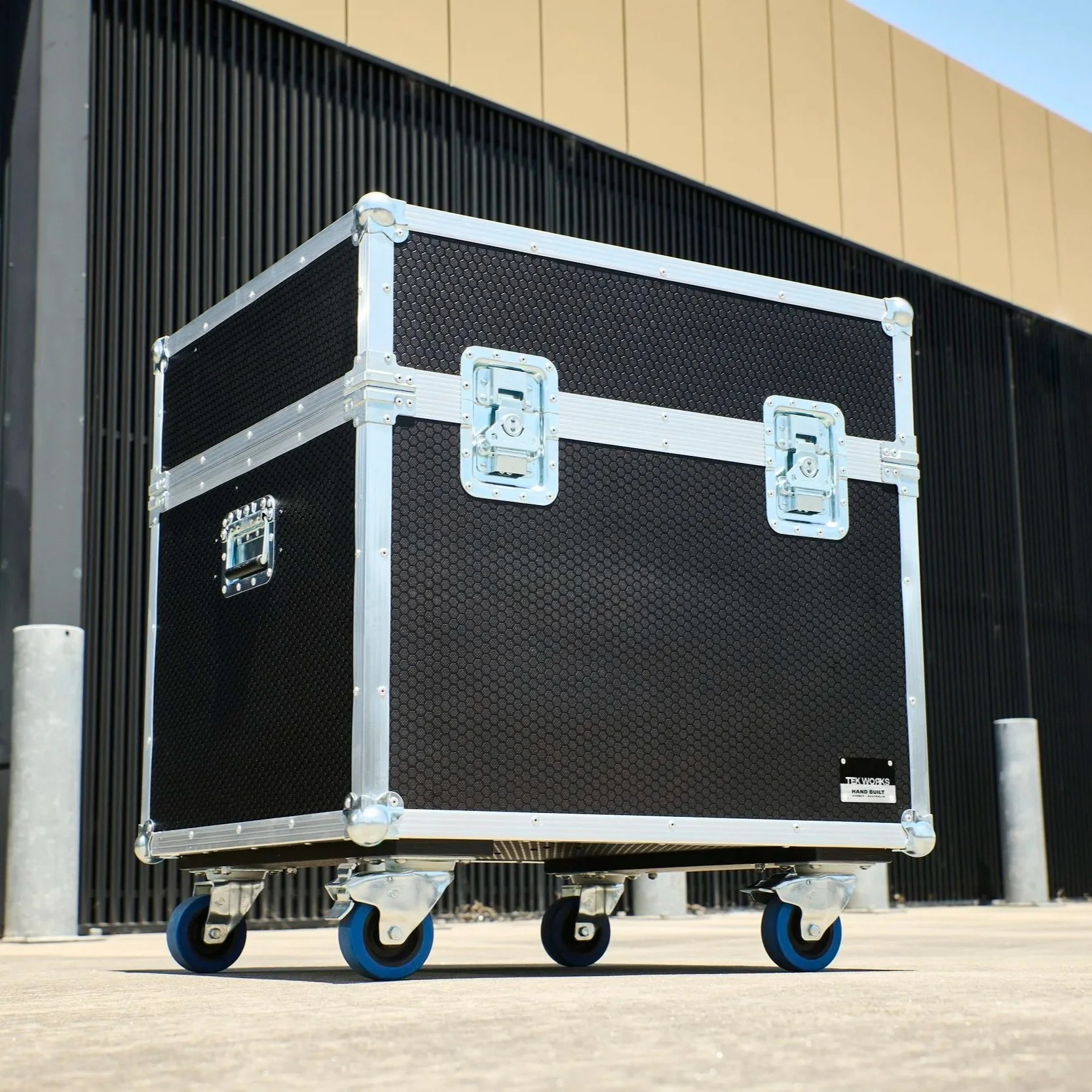 A black rolling equipment case with metal edges and handles, placed outdoors on a concrete surface in front of a black and beige building wall.