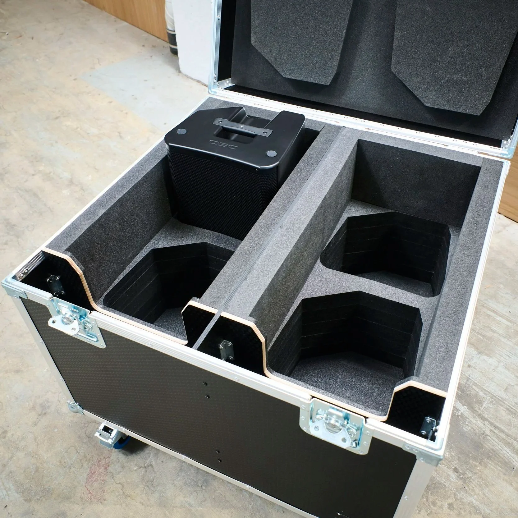 An open black equipment case with foam padding, containing a black speaker and compartments for other audio gear.