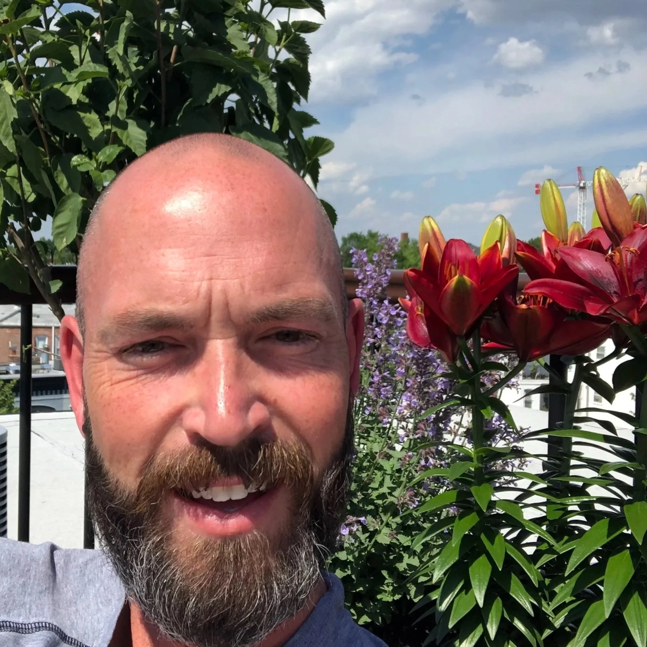 A man with a beard and shaved head smiling outdoors, surrounded by red and purple flowers with green foliage and a partly cloudy sky in the background.