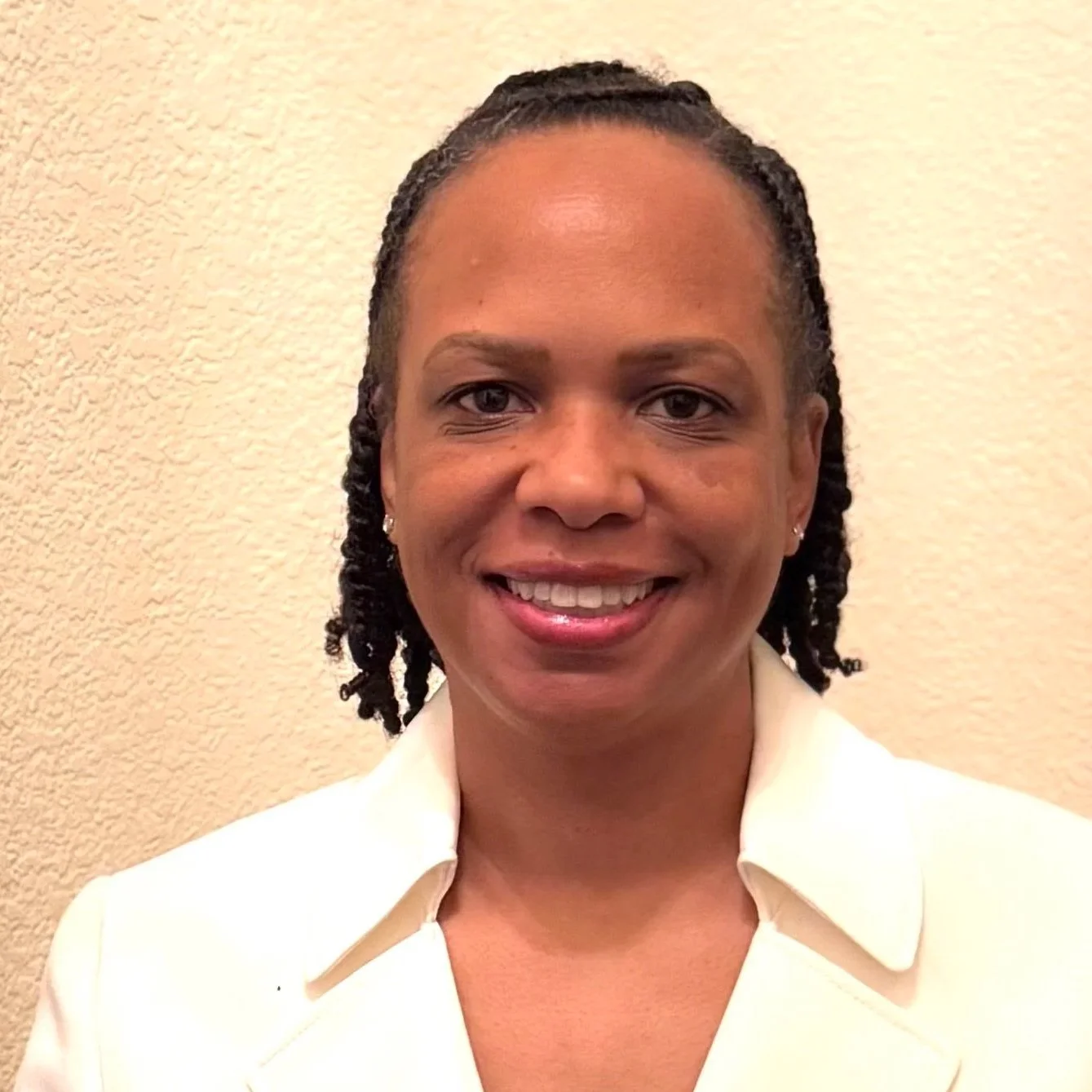 Close-up of a smiling woman with short, twisted hairstyle, wearing a white blazer in front of a beige textured wall.