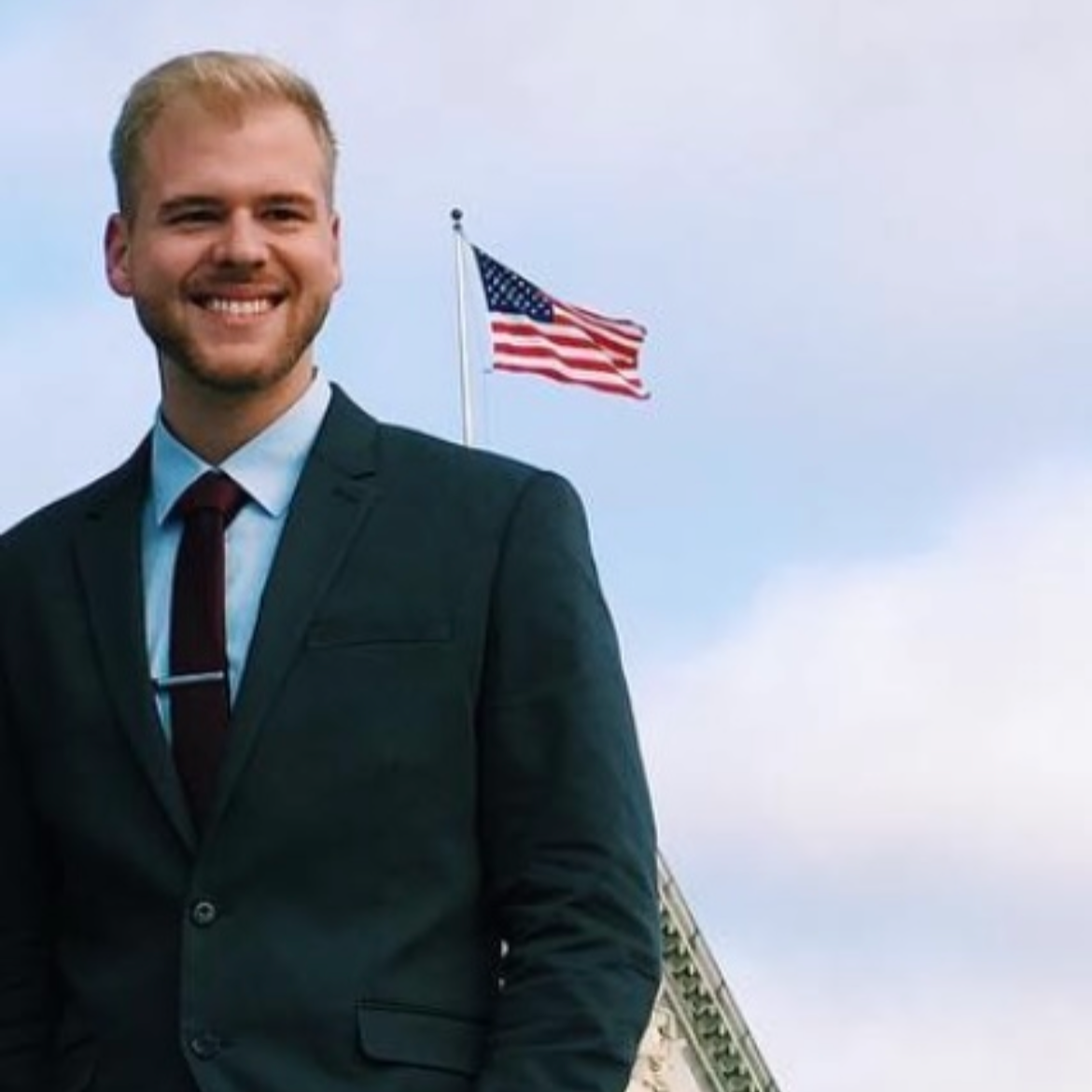 A young man wearing a suit and tie, smiling, standing outdoors with an American flag waving in the background.