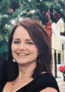 A woman with brown hair smiling outdoors, with flowers and a building in the background.