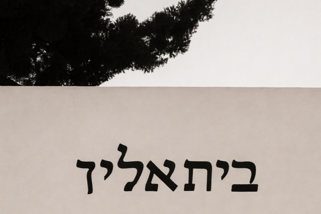 Close-up of a sign with Hebrew text on a beige wall, with part of a tree and the sky visible above.