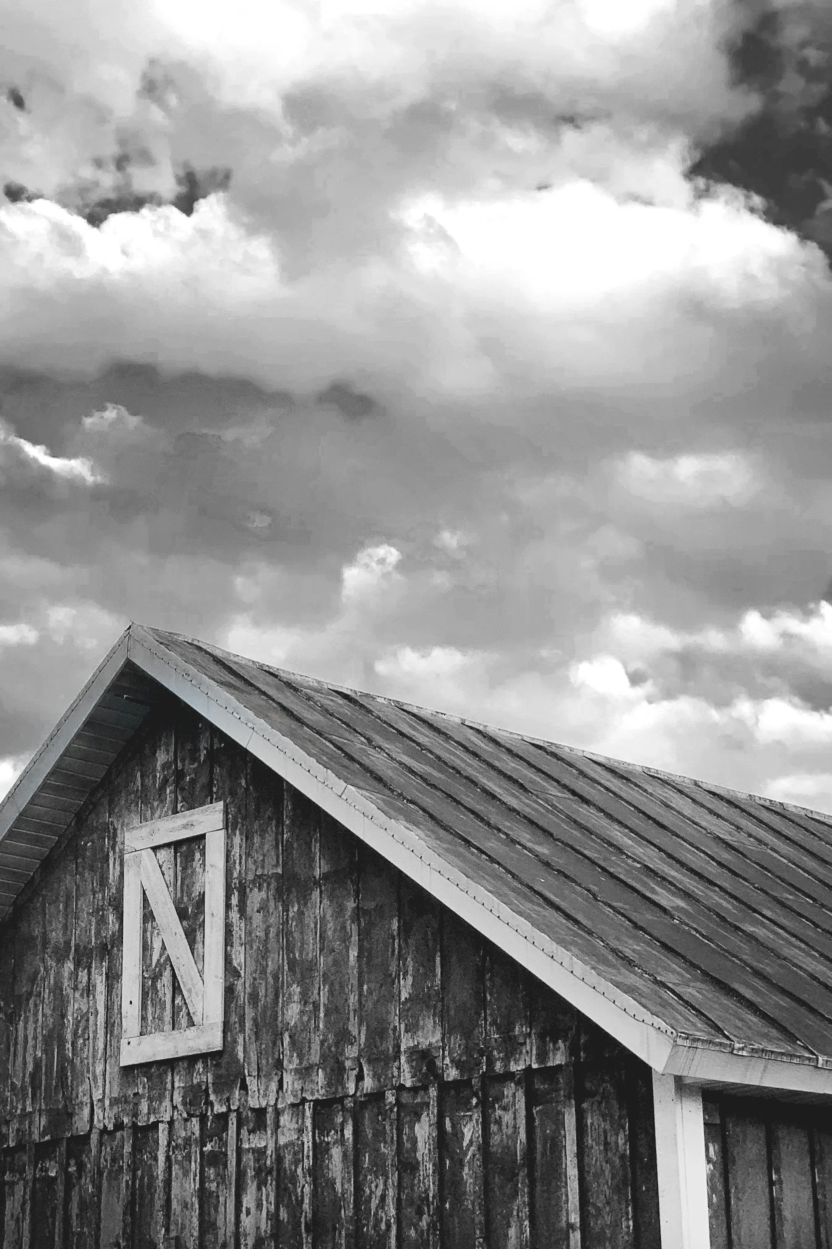 Black and white photo of a rustic wooden barn with a sloped metal roof, partly covered with moss or weathered wood, against a sky filled with cloudy weather.