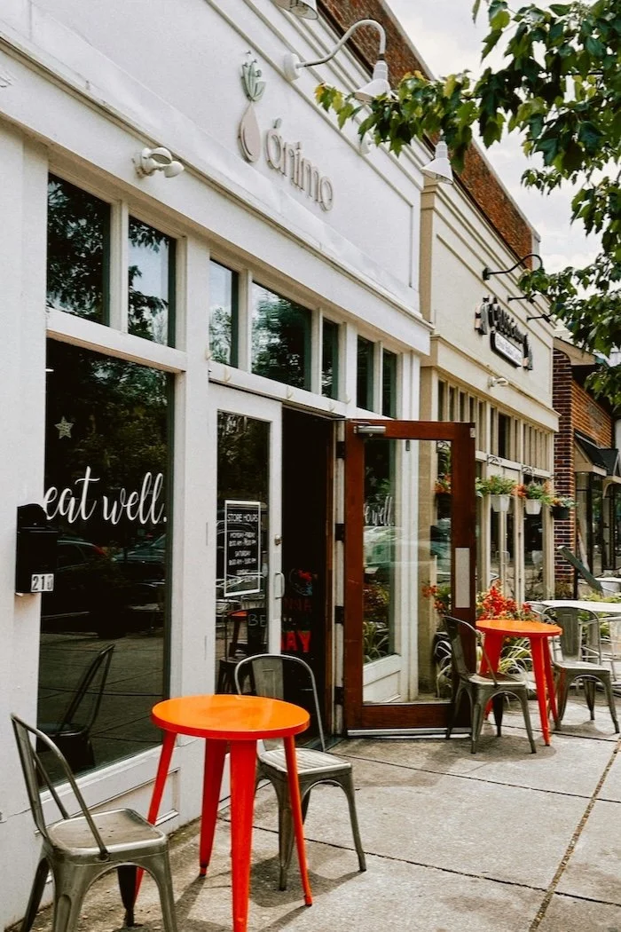 Street view of a storefront with outdoor seating, orange tables, black chairs, large windows, and signs for a restaurant called "Yum" and another sign reading "eat well."