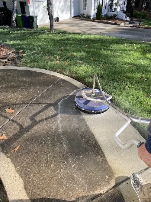 Person using a power washer on a sidewalk in front of a house with a lawn and driveway.