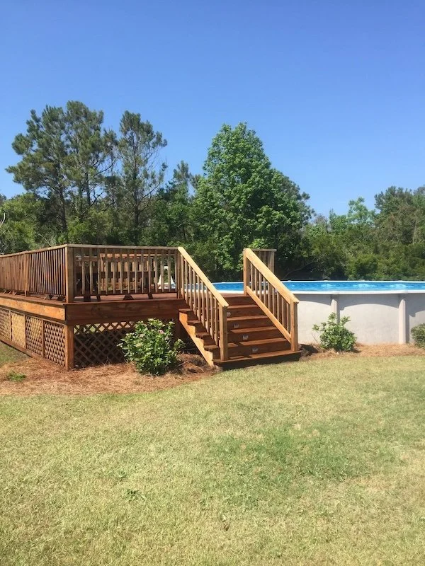 Above ground swimming pool with a wooden deck and stairs, surrounded by green grass and trees under a clear blue sky.
