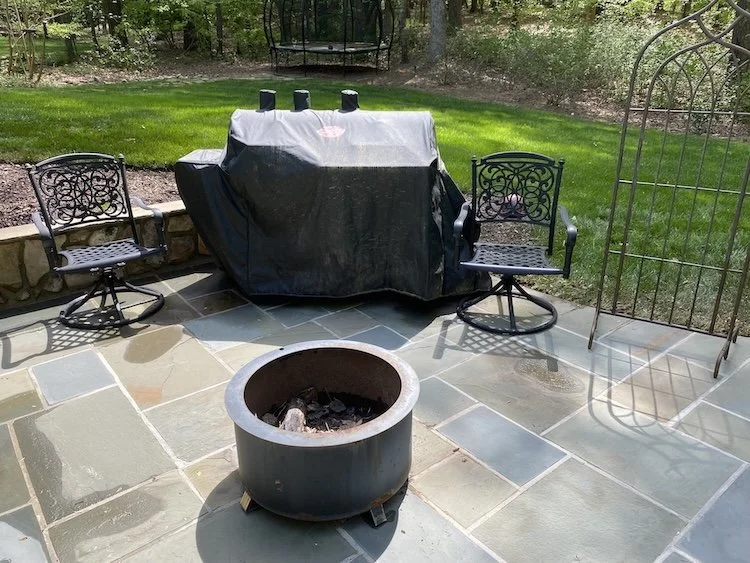 Patio with two metal chairs, a round fire pit, a covered barbecue grill, and a garden with green grass and trees in the background.