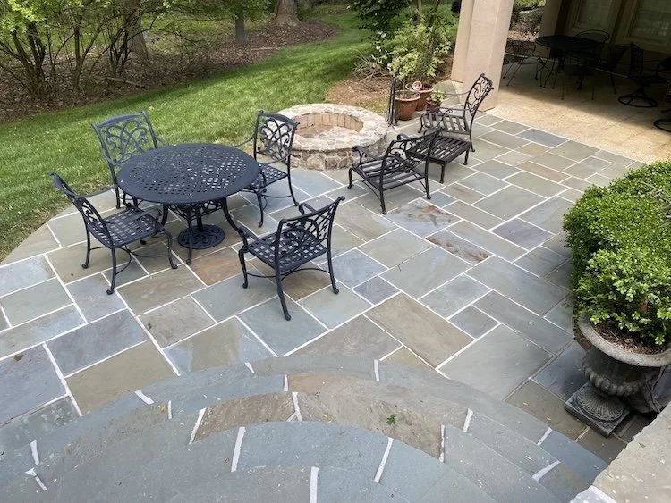 A backyard patio with a round black metal table and four matching chairs, a fire pit, and a stone paver floor. There are plants and bushes around the edges, and part of the house's patio with additional seating is visible in the background.