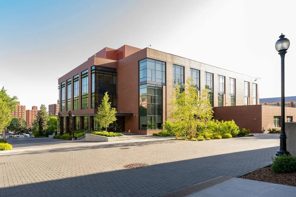 Modern brick building with large glass windows and surrounding greenery, under a clear sky.