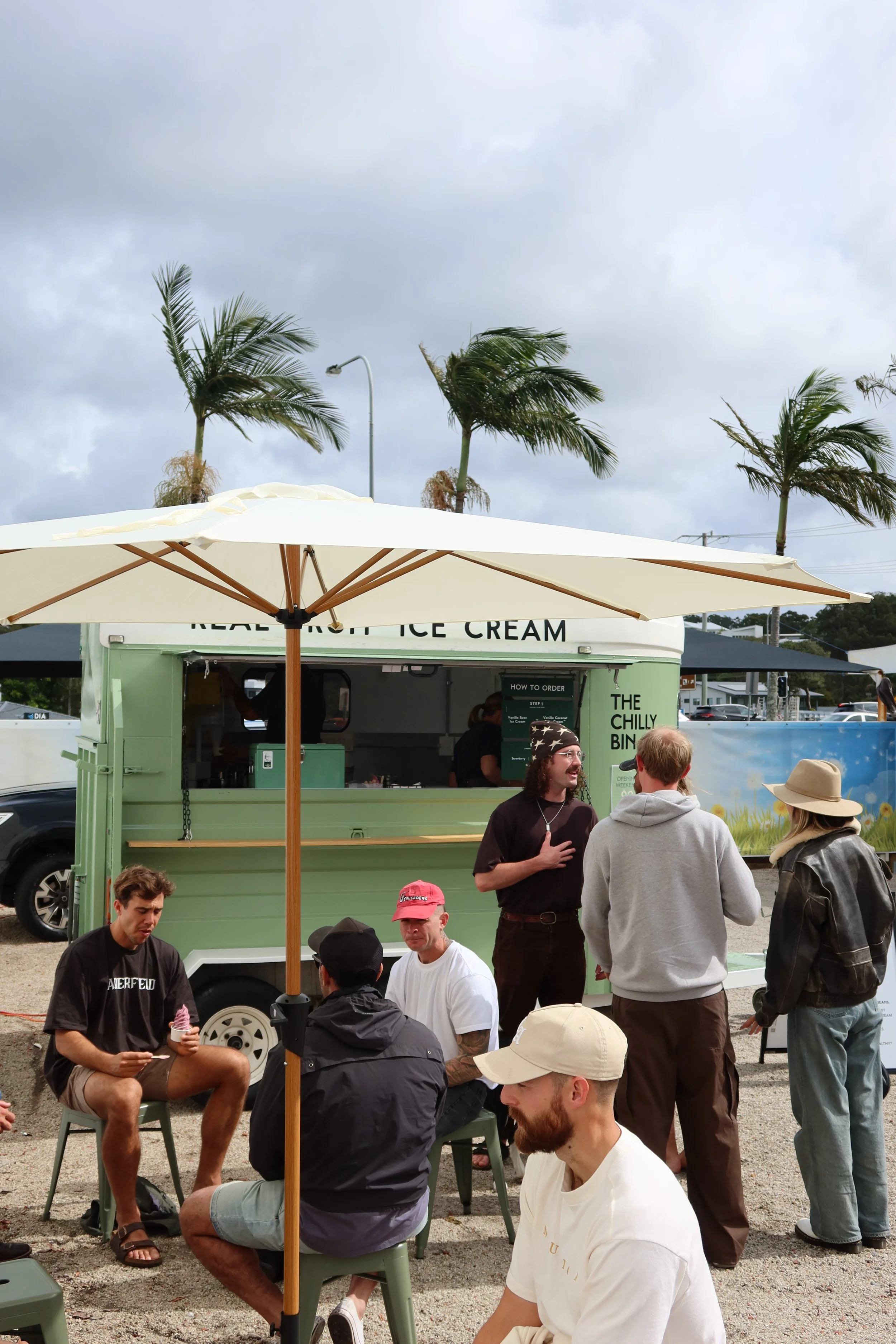 A group of people at an outdoor ice cream stand with palm trees and a cloudy sky in the background.