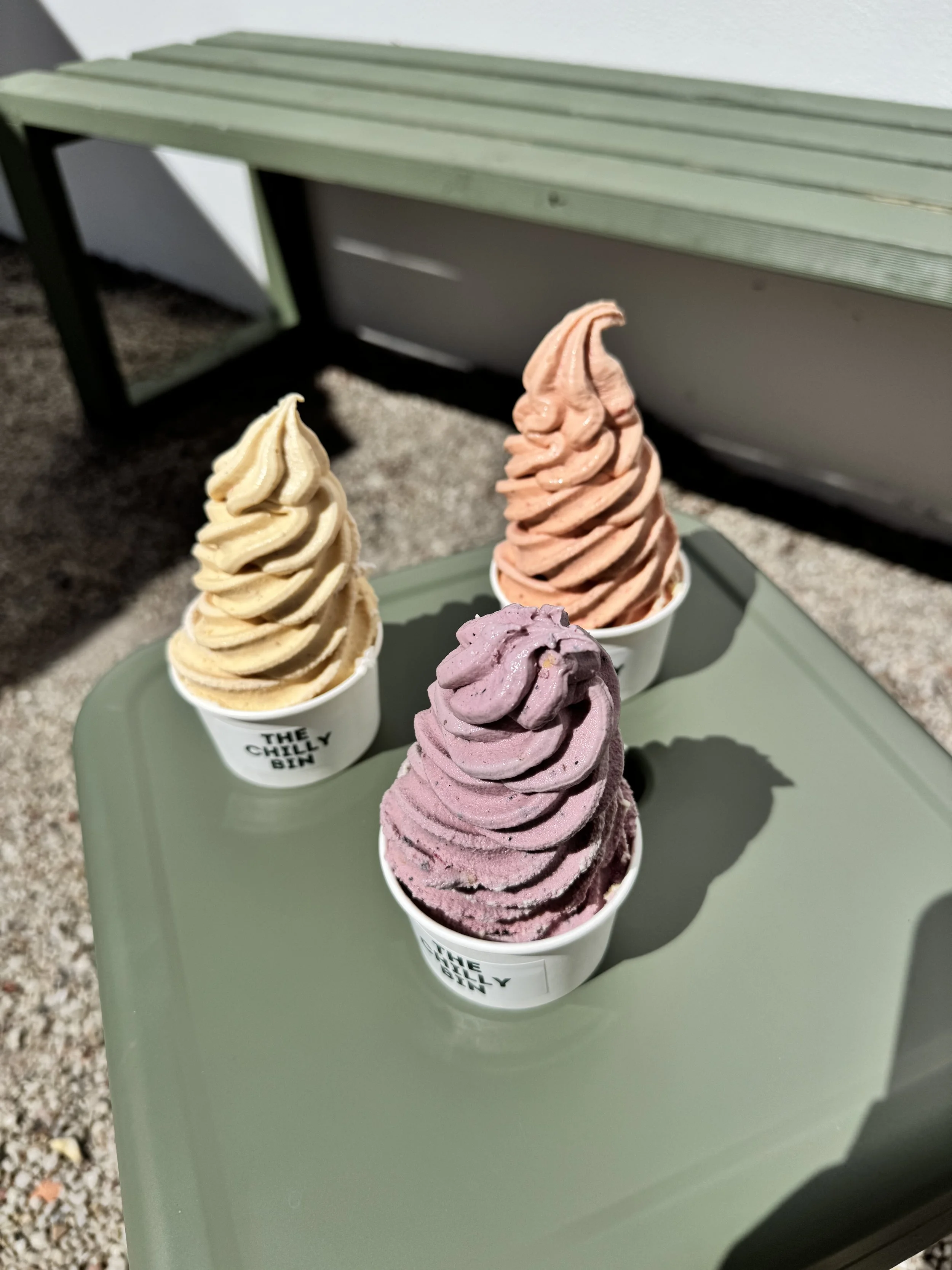 Three cups of colorful soft-serve ice cream on a green table outdoors, with a bench and gravel in the background.