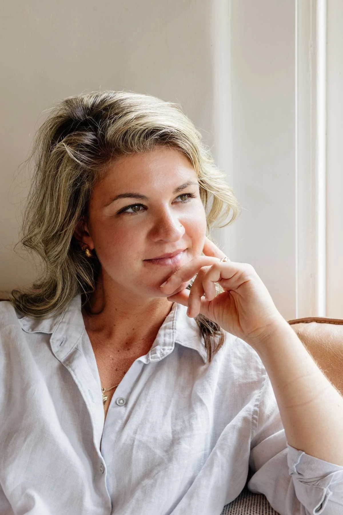 A woman, Nikki Williams with blonde hair and light skin sitting near a window, wearing a white shirt and gold jewelry, resting her chin on her hand, looking thoughtfully out of the window.