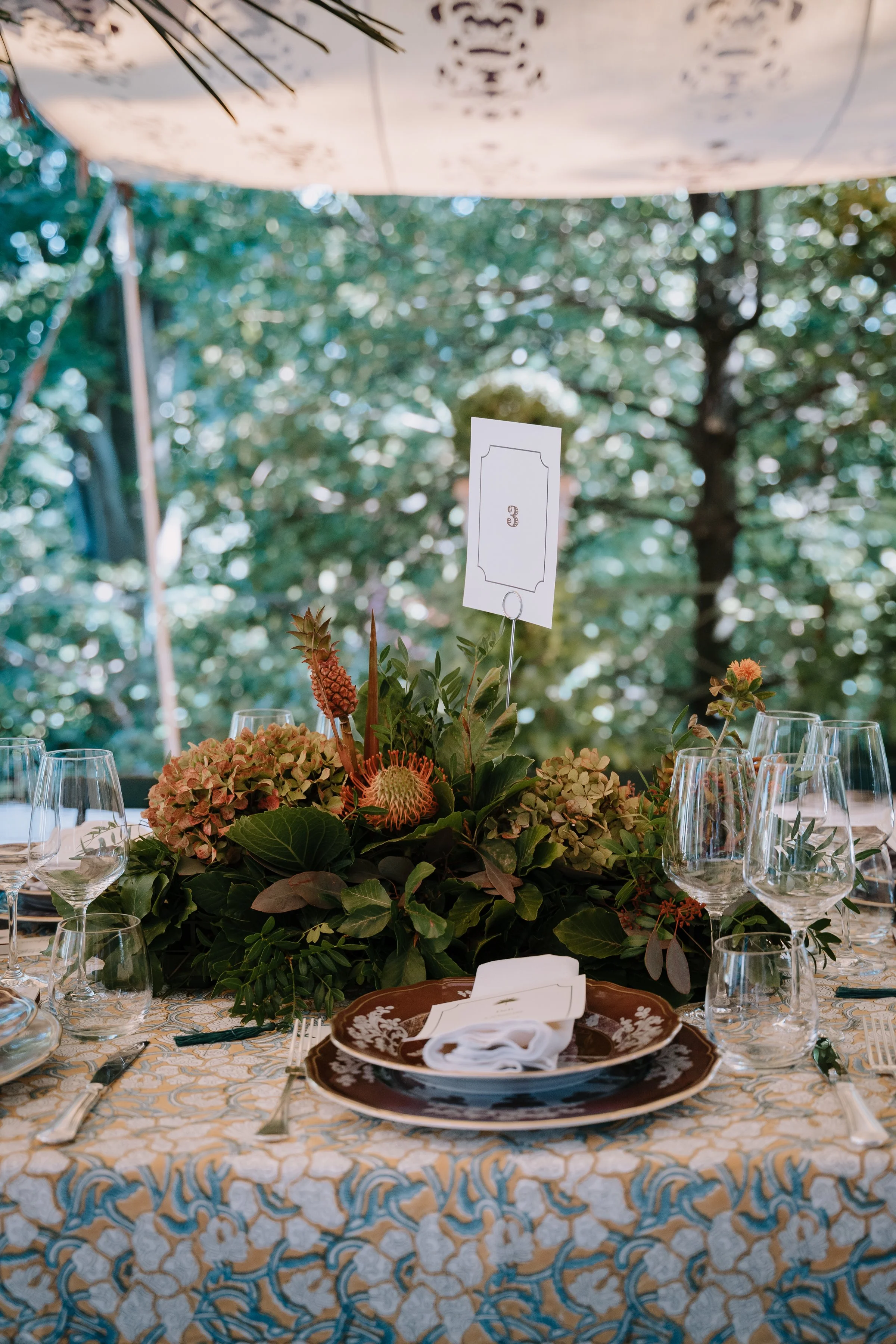 Tavola apparecchiata con piatti bianchi, bicchieri di vetro e posate, decorata con un centro tavola di fiori e foglie verdi, sotto una tenda con decorazioni floreali. Segnaposto con il numero 3.