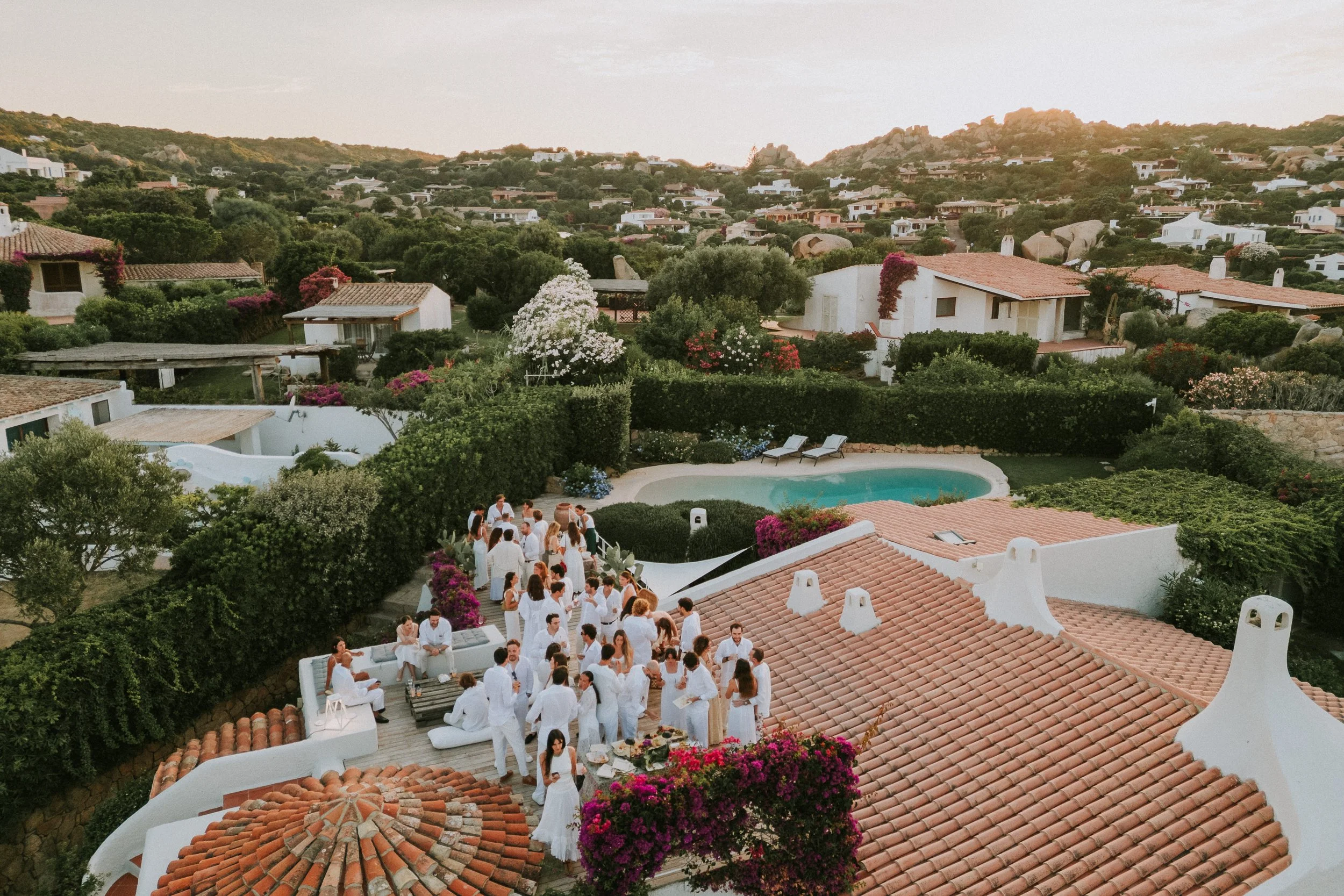 Gruppo di persone vestite di bianco che socializzano su una terrazza con vista su case e un giardino con piscina, in un paesaggio collinare in Sardegna, Porto Rafael.