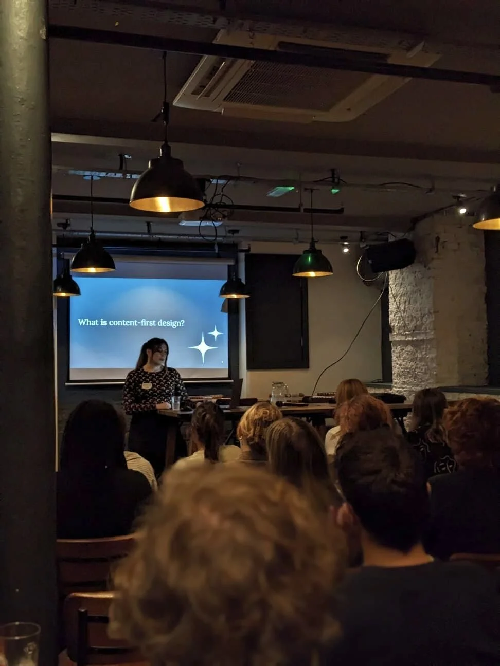 Me, a white woman in her late 20s, presenting at a UX meet-up. I'm looking at a laptop for notes, while the audience watches. Behind me is a screen that says "What is content-first design?"