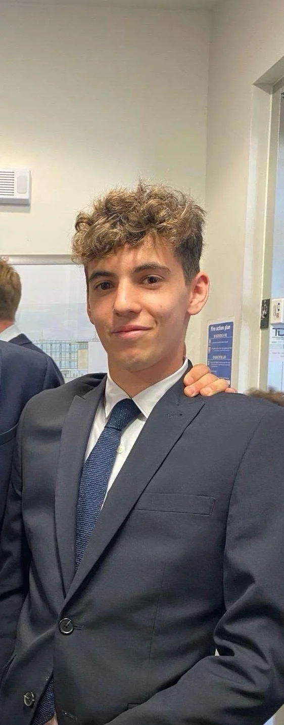 A man with curly dark hair, glasses, a nose ring, and a beard, wearing a dark green sweater and a white shirt underneath, is sitting indoors with a neutral expression, looking slightly to the side. The background features a blurred plant and wall.