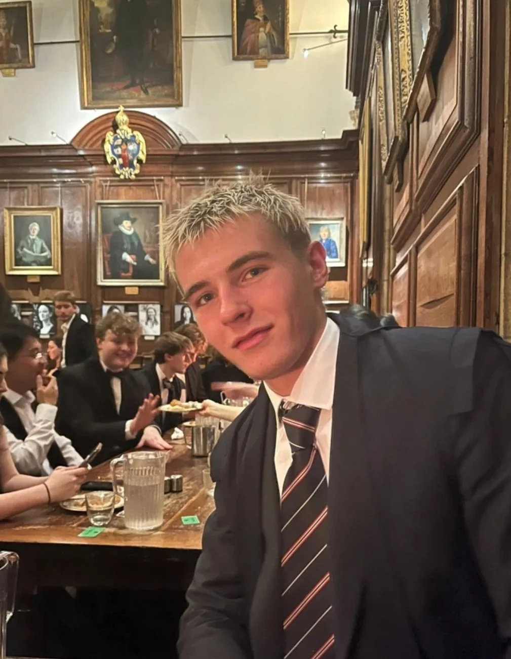 Young man with blonde hair wearing a black suit, white shirt, and striped tie, sitting at a wooden table in a historic room with wood-paneled walls, surrounded by other people in formal attire, with portraits and a crest hanging on the wall behind him.