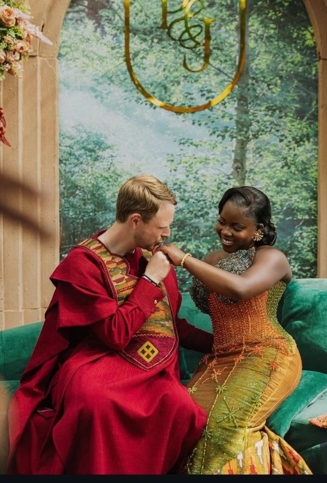 Een man en een vrouw die lachen en een ring uitwisselen, zittend op een groene bank met een bosachtige achtergrond en een decoratief gouden monogram boven hen.