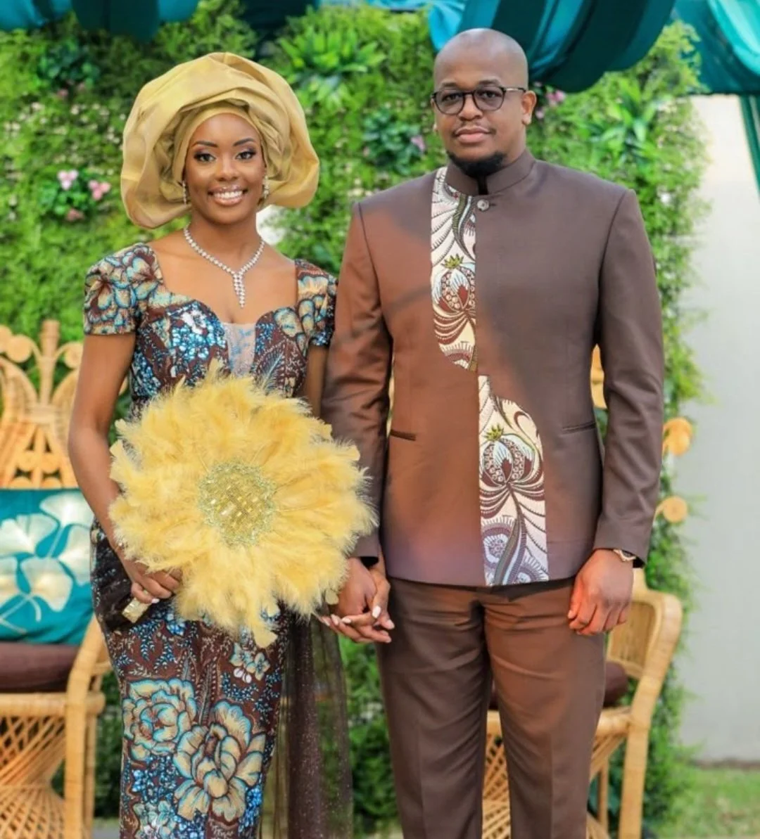Een man en vrouw in traditionele en formele kleding, hand in hand, op een decoratieve achtergrond van groen en bloemen, mogelijk tijdens een speciale gelegenheid of ceremonie.