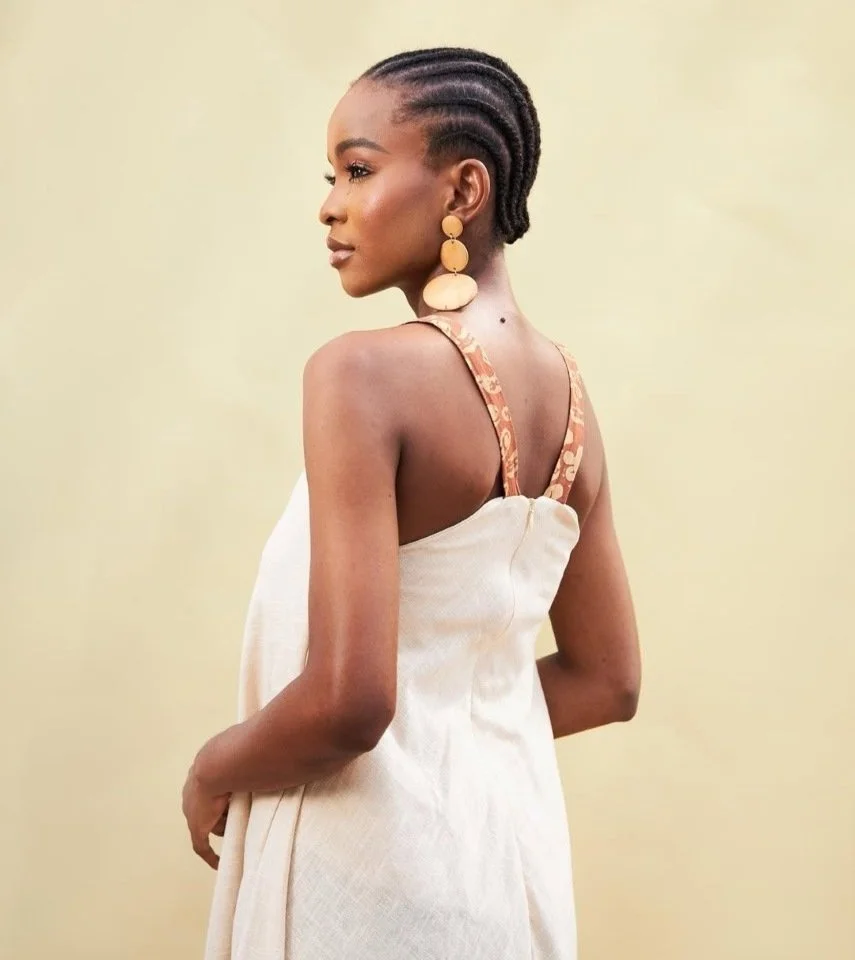 Een jonge vrouw met gevlochten haar die een wit jurkje draagt met bandjes in een bruin patroon, staand tegen een lichte achtergrond.