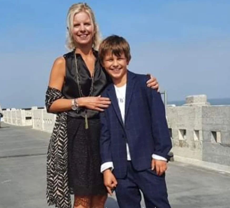 Een vrouw en een jongen poseren samen buiten, ze lachen en hebben elkaar omarmd. De vrouw draagt een zwart en wit gekleed, de jongen draagt een blauwe pak. In de achtergrond is een muur en een heldere blauwe lucht te zien.