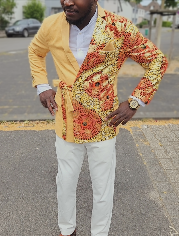 Man draagt een gestileerd jack met een opvallend kleurrijk patroon en een gele kleur, witte blouse en witte broek, en draagt een horloge.