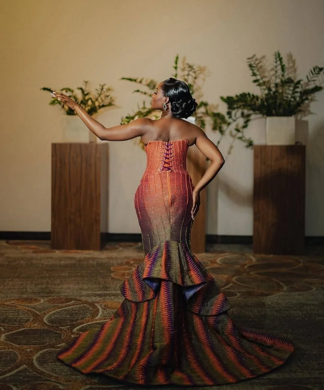 Een vrouw in een opvallende, kleurrijke avondjurk poseert binnen, met haar arm uitgestrekt en haar hoofd schuin.