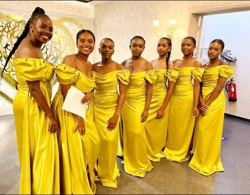 Zeven vrouwen in gele galajurken staan samen in een kamer, lachend en poserend voor de camera.