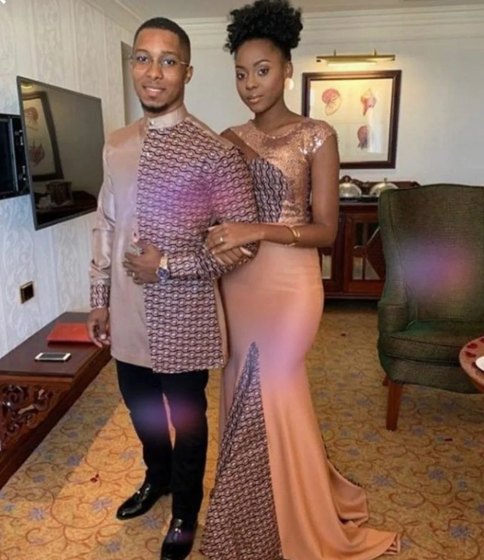 Een man en een vrouw staan samen in een woonkamer, beiden gekleed in traditionele, elegante kleding met patronen en glitters. Ze kijken naar de camera en zijn gekleed voor een speciale gelegenheid.
