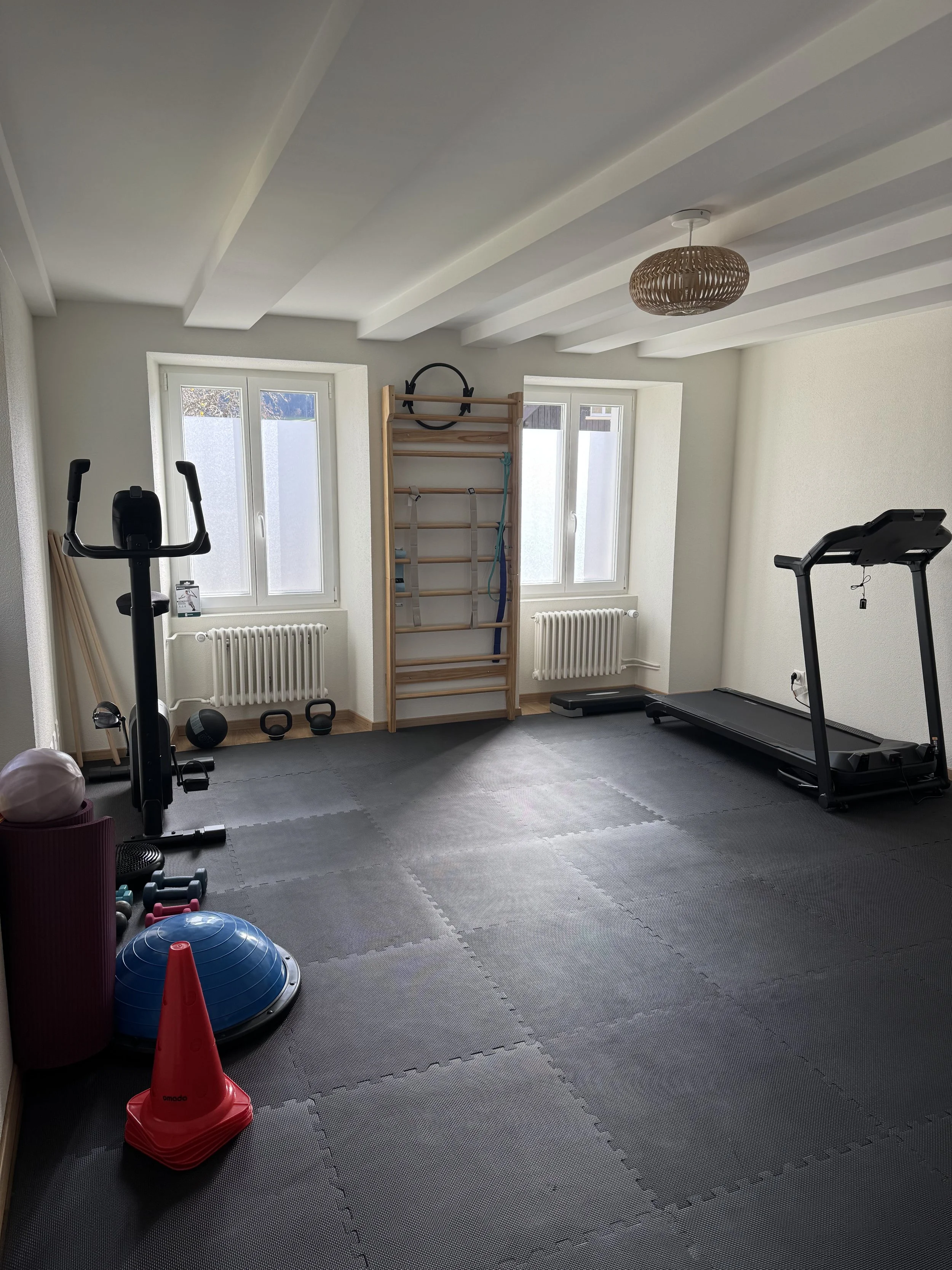 Salle de sport avec tapis de course, ballons de fitness, cônes de signalisation, matériel d'entraînement, fenêtres laissant entrer la lumière naturelle, sol en tapis gris, luminaires au plafond, mur clair.