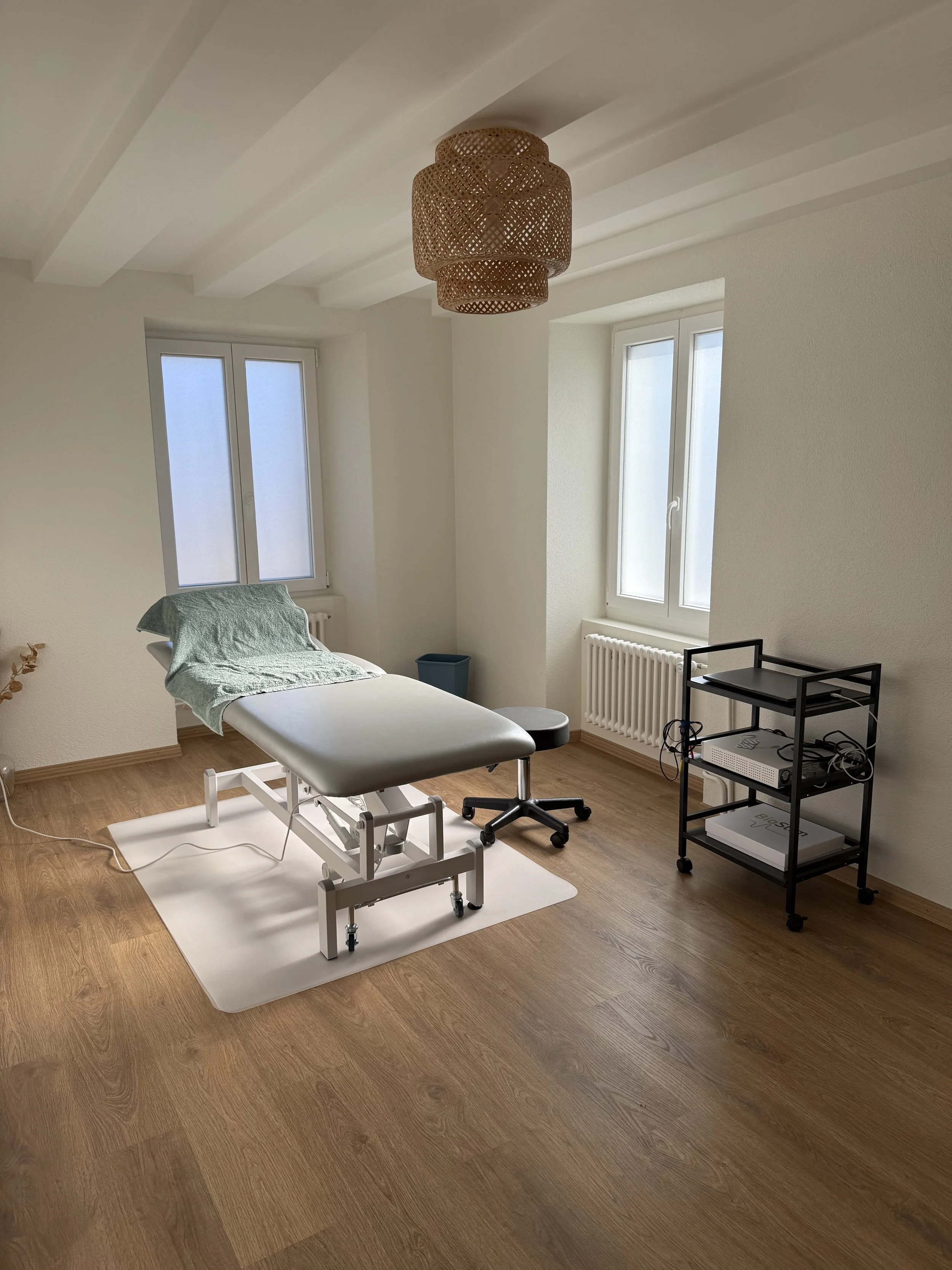 Une salle de traitement ou de consultation médicale avec une table d'examen, deux fenêtres et un éclairage suspendu en rotin.