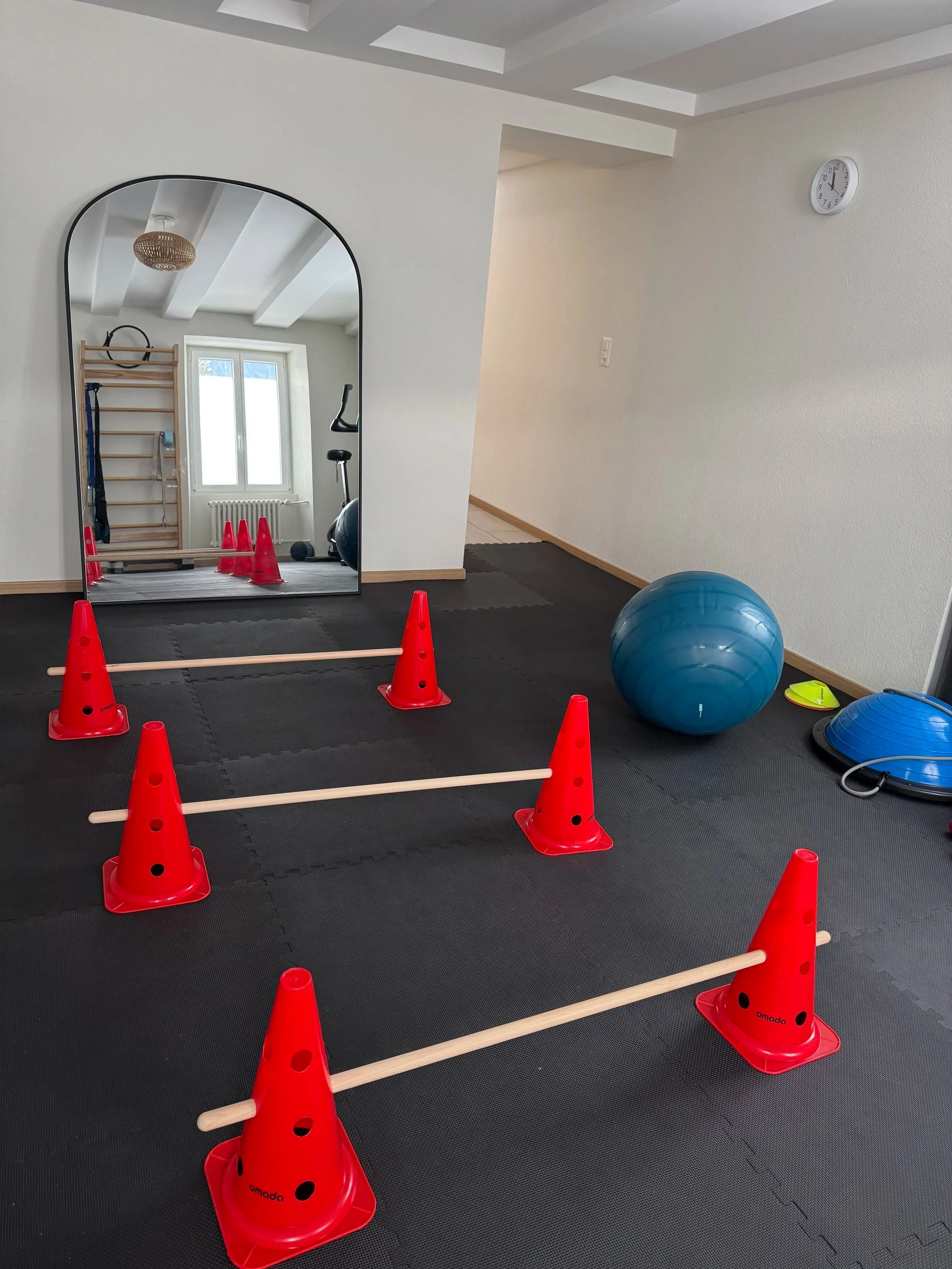 Une salle d'entraînement équipée de matériel de fitness et d'exercices, avec des cônes orange, un ballon bleu, un tapis de sol noir, un miroir en arc, une échelle de gymnastique en bois, et un espace de gym dans une pièce lumineuse.