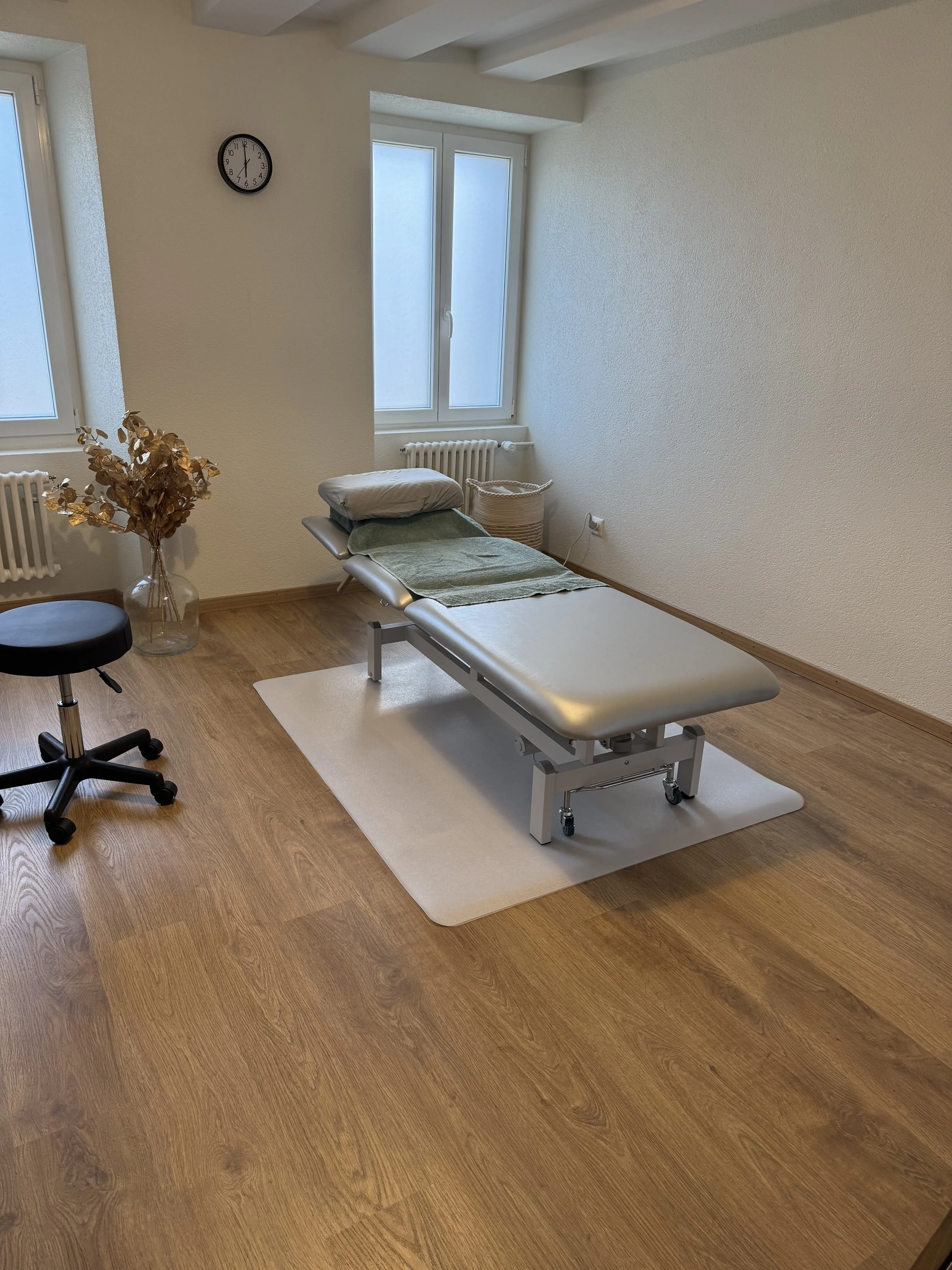 Salle de consultation médicale avec un lit d'examen, une plante en vase, un fauteuil noir, un mur blanc, une fenêtre, un radiateur, une horloge et un panier en osier.