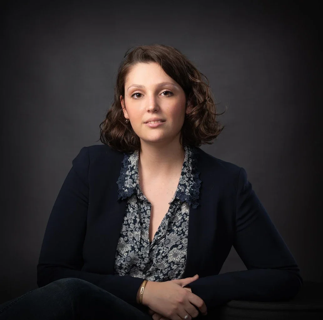 Portrait d'une femme avec des cheveux bruns, portant une veste noire et un chemisier floral, assise contre un fond sombre.
Cette femme est Maitre Lantillon-Ray.