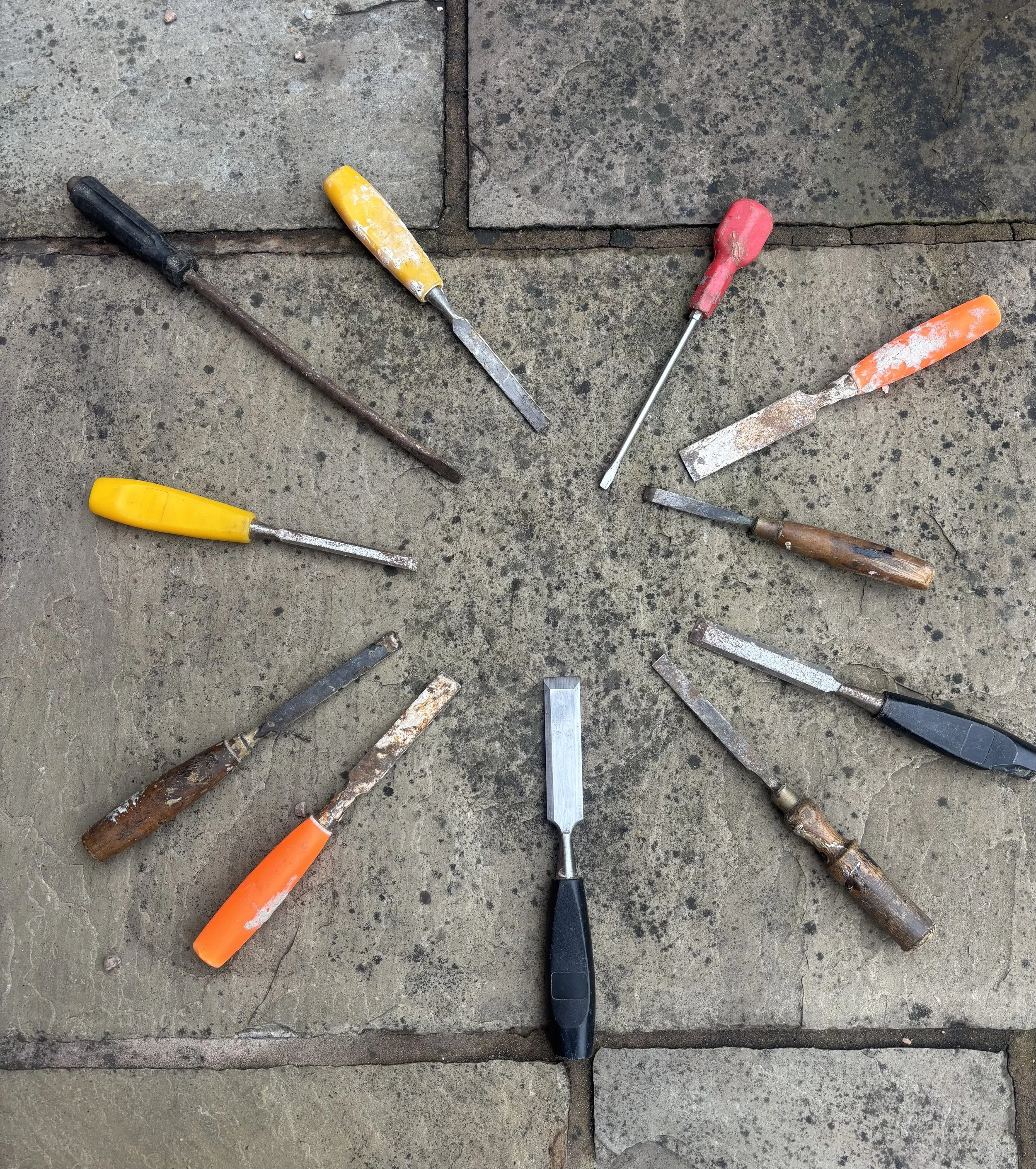 Collection of various hand chisels and carving tools arranged in a circle on a concrete surface.