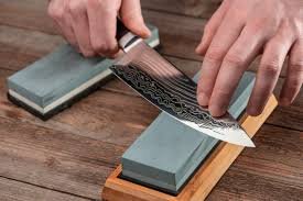 Person sharpening a knife using a whetstone on a wooden work surface.