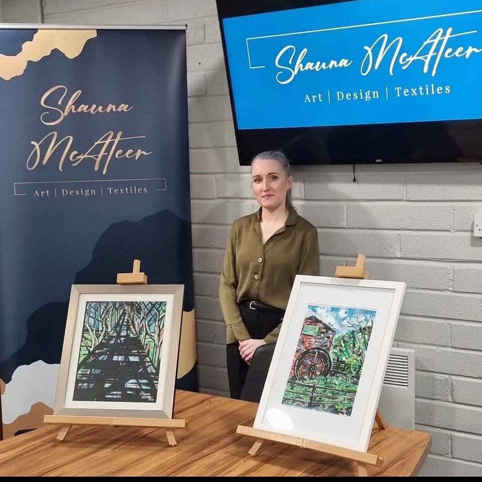 A woman standing behind a wooden table with two framed watercolor paintings on easels. One painting depicts a bridge or path through trees; the other shows a wagon in a rural landscape. Behind her is a blue banner with her name, Shauna McAfeer, and the words Art, Design, Textiles, on the left, and a screen displaying her name and services on the right.