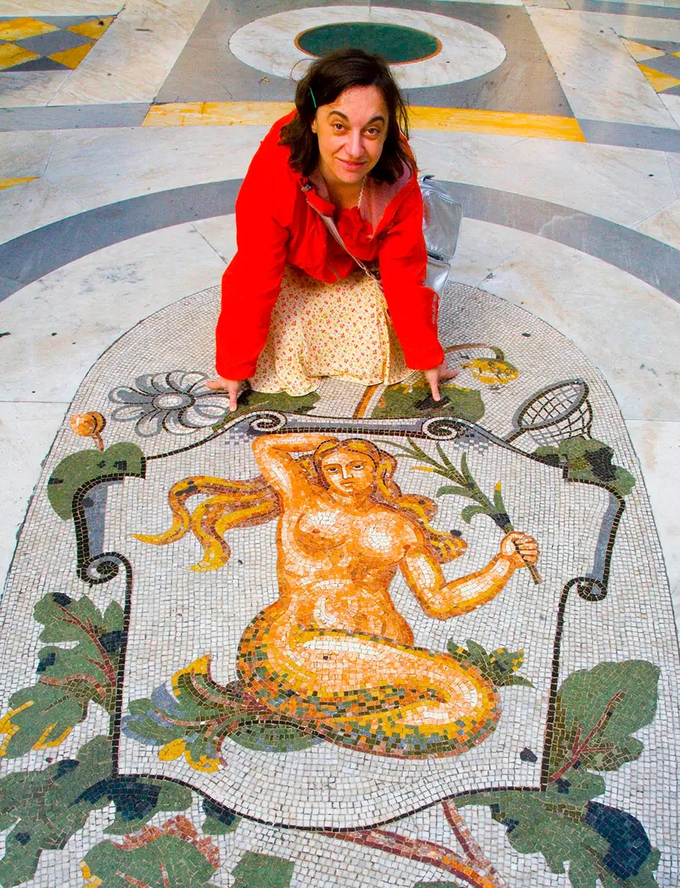 Portrait of the artist Aleksandra Mir sitting down next to an antique mosaic of a mermaid surrounded by green foliage. the artist is wearing a neon orange wind jacket and a feminine yellow skirt with small flowers on it. She is smiling.