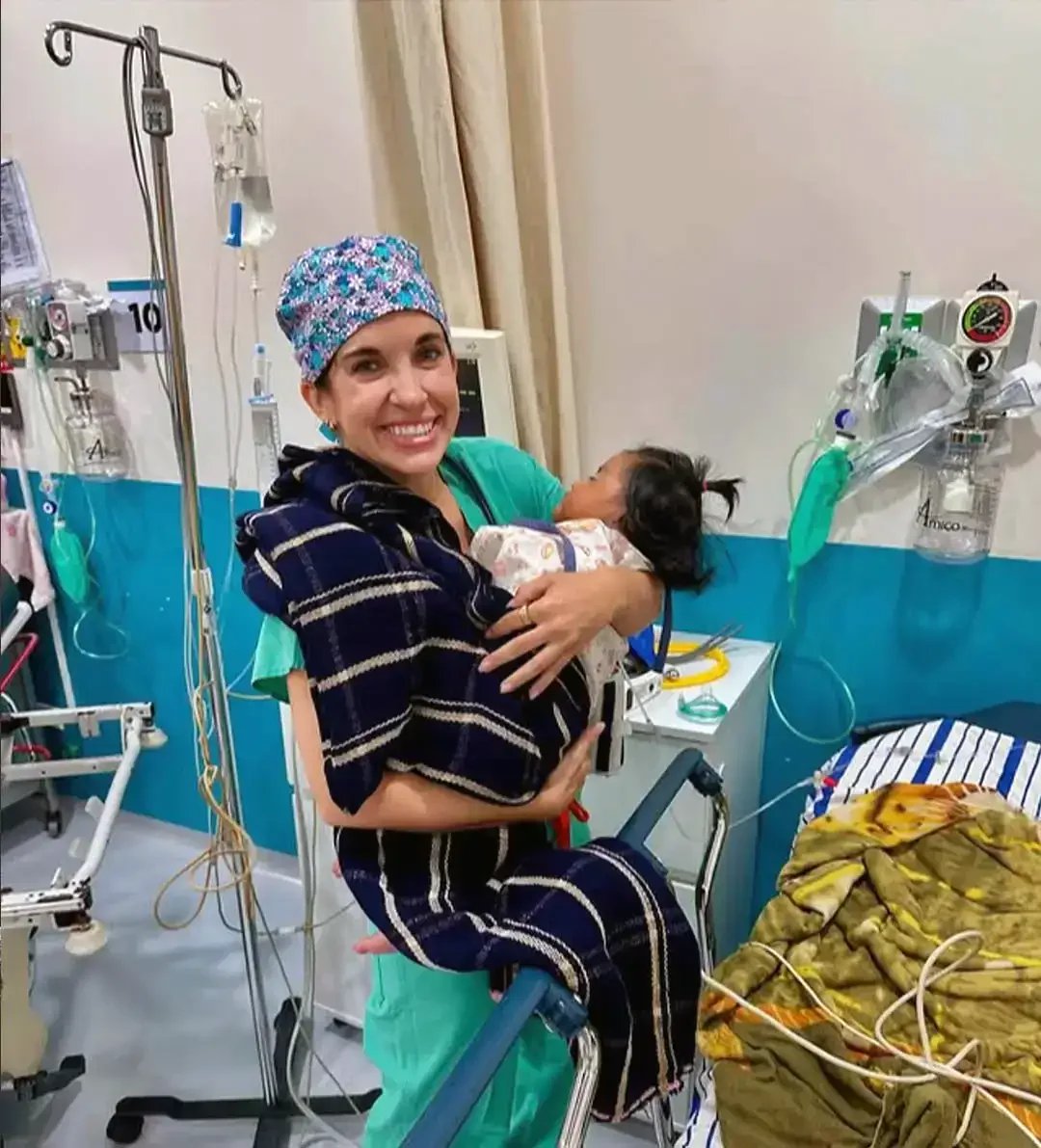A photograph of nurse practitioner Erin Kesler dressed in hospital scrubs holding a small baby surrounded by medical furniture and equipment