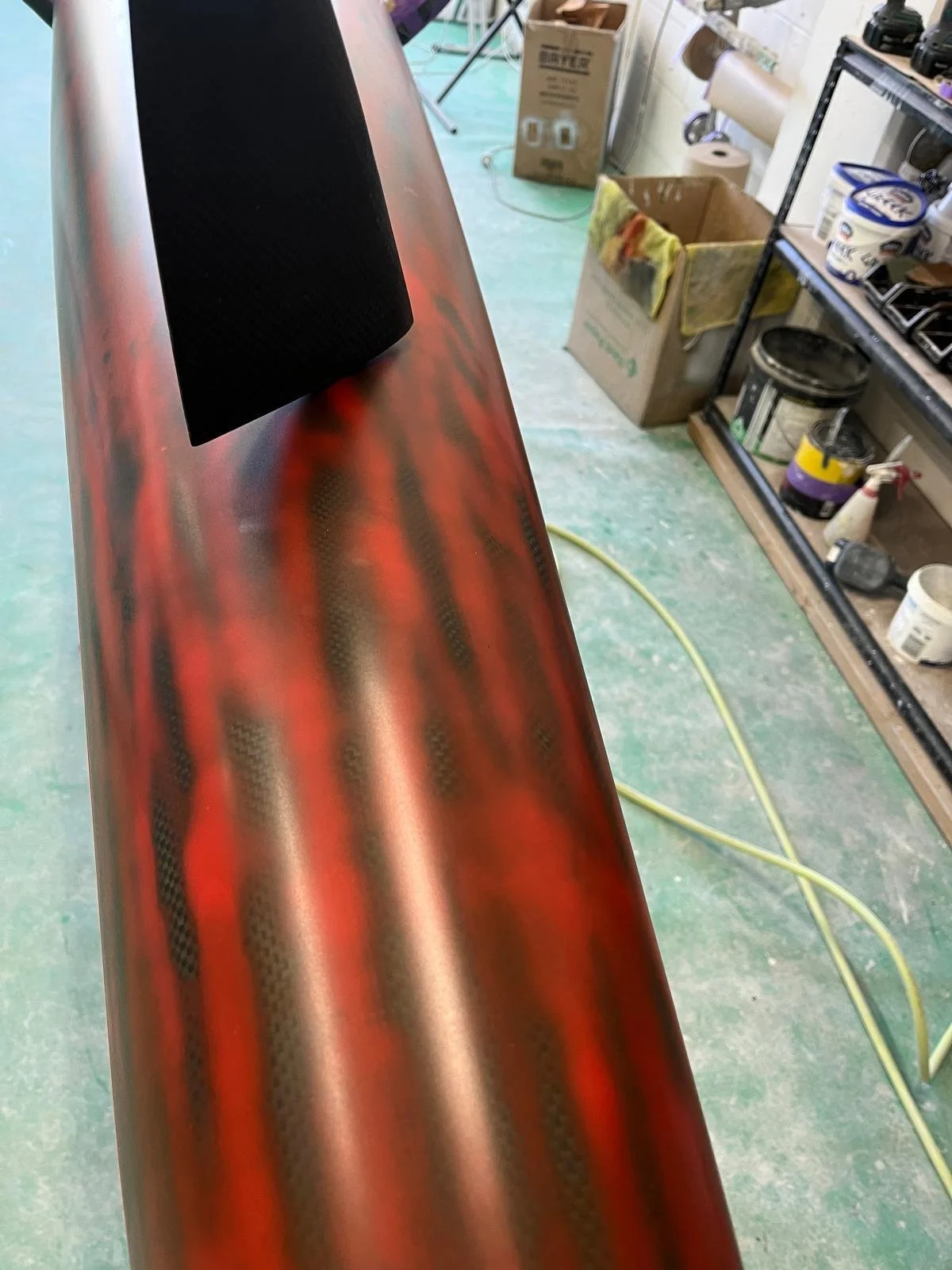 Close-up of a red surfboard with a black fin, inside a workshop with various tools, supplies, and cardboard boxes in the background.