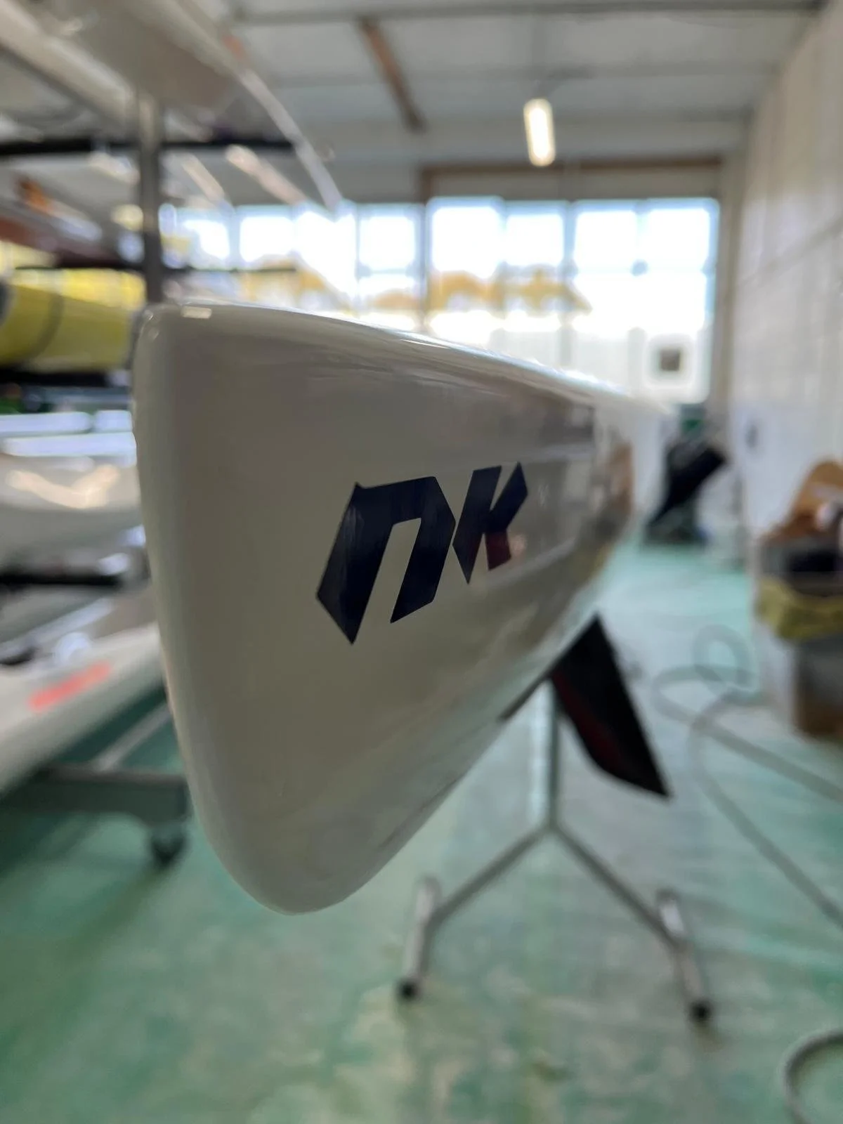 Close-up of a white surfboard with the logo 'DK' in black and red, in a warehouse or storage area with boat equipment and shelves in the background.