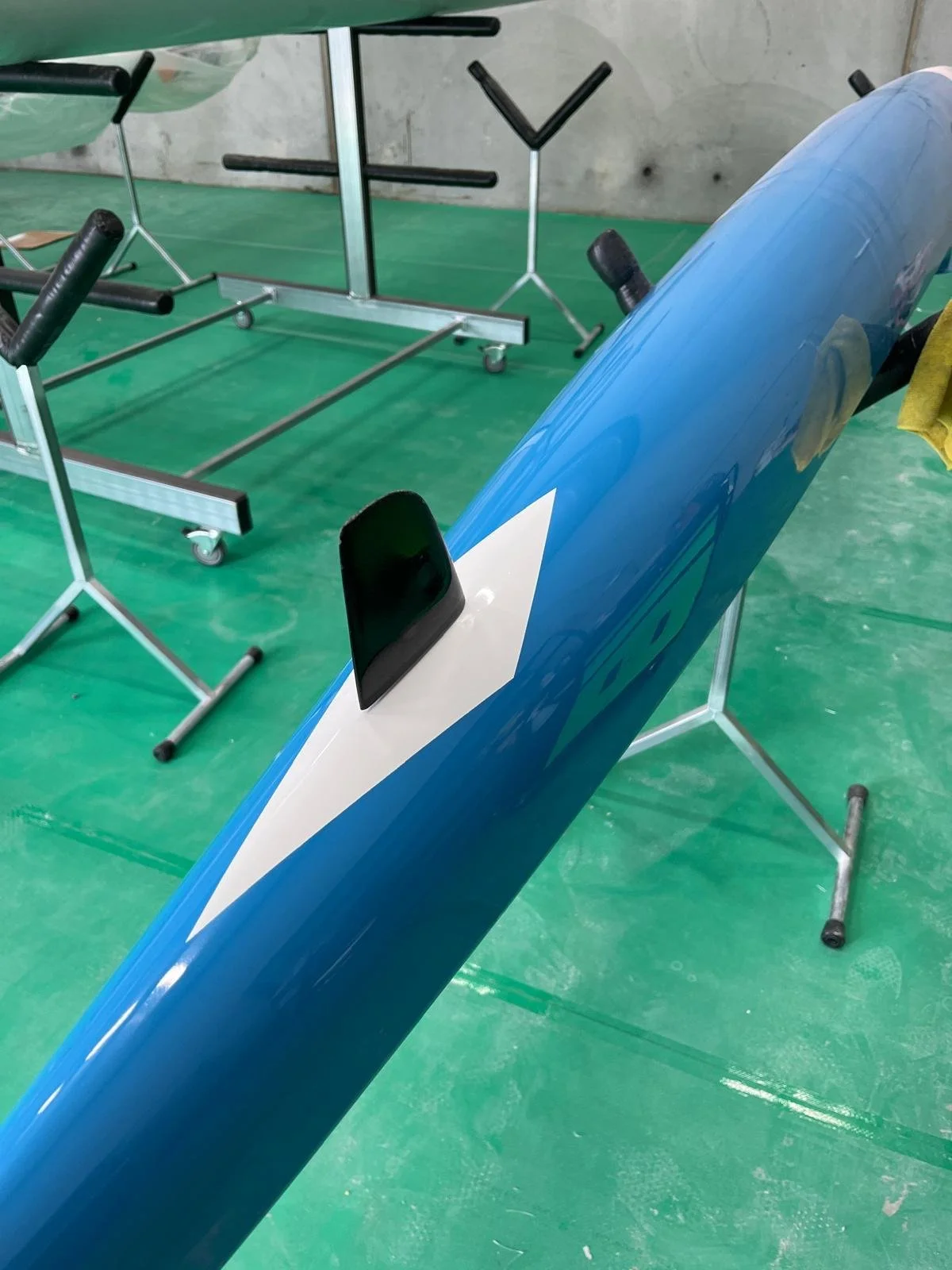 Close-up of a blue and white kayak with a black fin, placed on a stand in a workshop or storage area with green flooring and equipment in the background.
