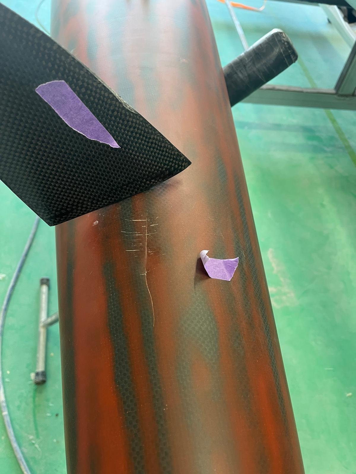 A close-up of a red and black carbon fiber bicycle frame with purple painter's tape on it, in a workshop setting.