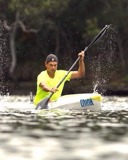 Person wearing a yellow shirt and black cap kayaking on water, using a paddle to navigate.