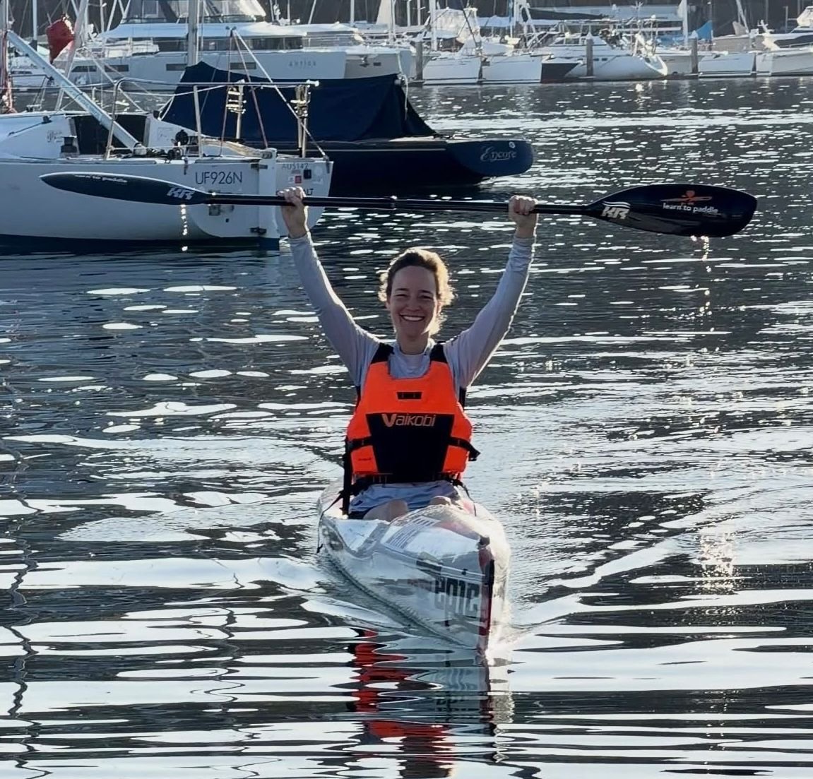 A woman smiling and celebrating while sitting in a kayak on a body of water, holding a paddle upright above her head. She is wearing an orange life vest and is surrounded by docked boats.