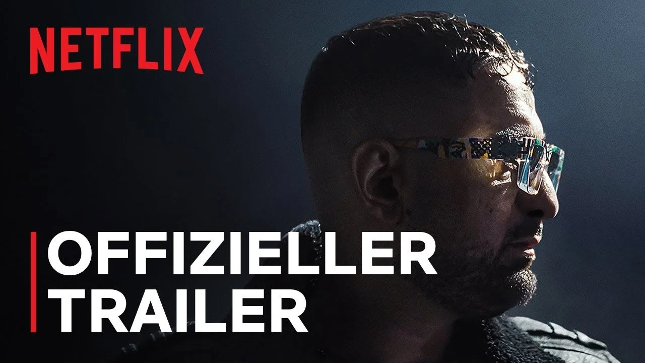 Das YouTube-Thumbnail der Doku. Oben links das Netflix-Logo, unten links "Offizieller Trailer" und rechts ein nach rechtsschauender Haftbefehl mit Sonnenbrille im Scheinwerferlicht.