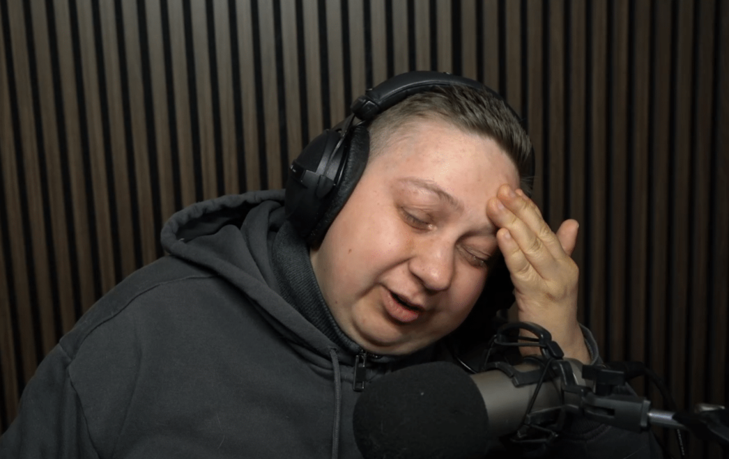 Timgioh sitzt traurig vor einer Holzwand und fasst sich an die Stirn.