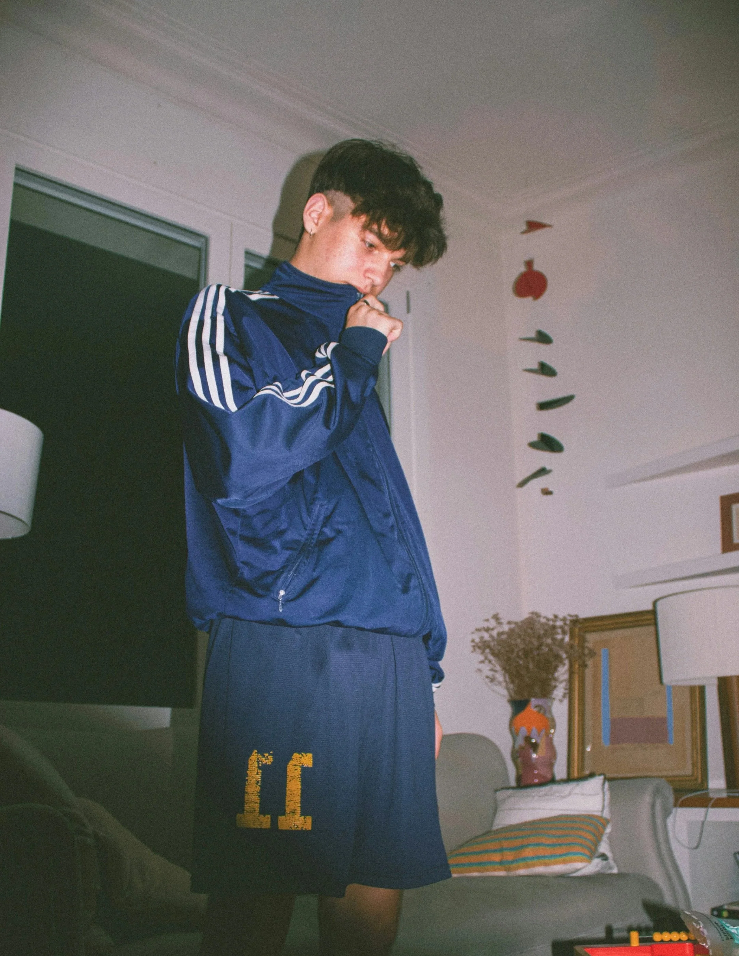 Junge in einem Zimmer mit Blitz fotografiert. Er hat eine blaue Adidas-Jacke und eine kurze, blaue Sporthose mit der Nummer 11 darauf an. Er schaut runter.